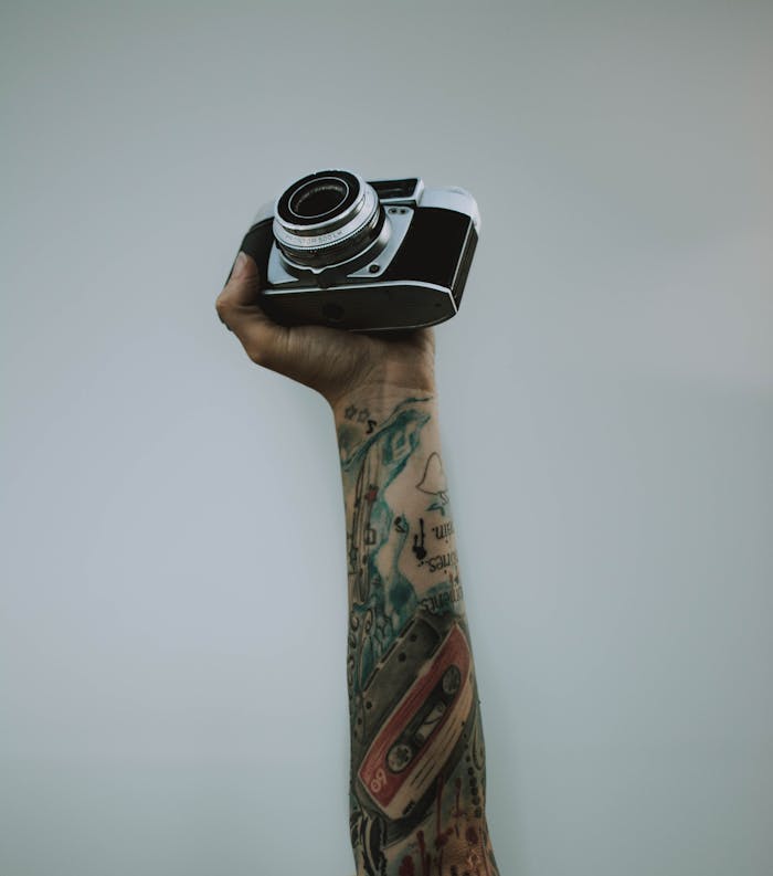 A tattooed arm raises a vintage camera against a plain backdrop, showcasing modern art and photography.